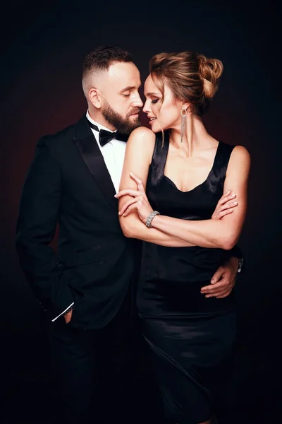 Casal luxuoso e elegante: homem barbudo bonito vestindo smoking com mulher incrível com cabelo loiro updo, vestindo vestido preto sedoso e jóias chiques, posando em estúdio escuro — Fotografia de Stock