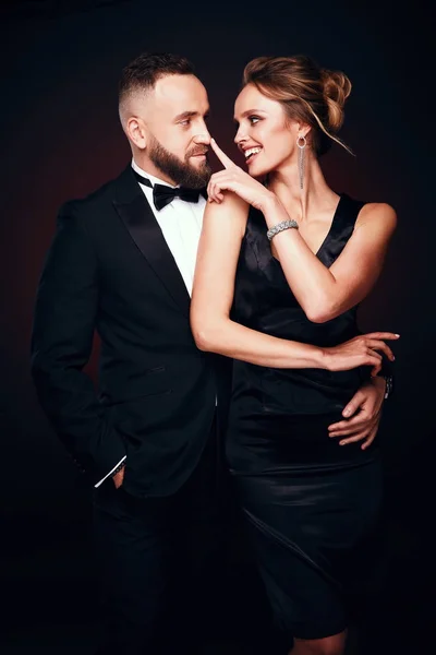 Happy and elegant couple: handsome bearded man in tuxedo with amazing woman with blonde updo hair, wearing silky black dress and chic jewelry, posing in dark studio