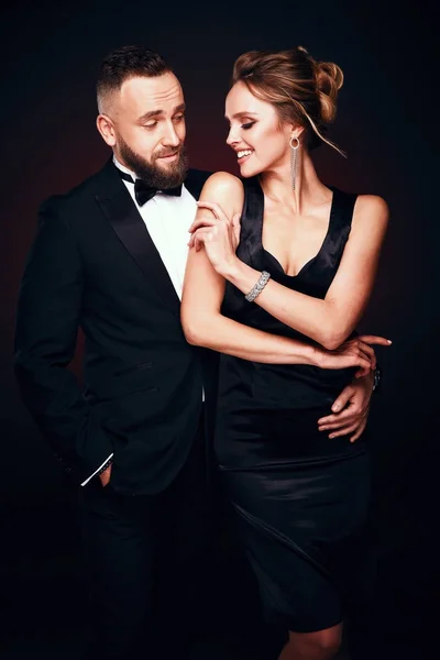 Casal de luxo: homem barbudo bonito em smoking com mulher incrível com cabelo loiro updo, vestindo vestido preto sedoso e jóias chiques, posando em estúdio escuro — Fotografia de Stock