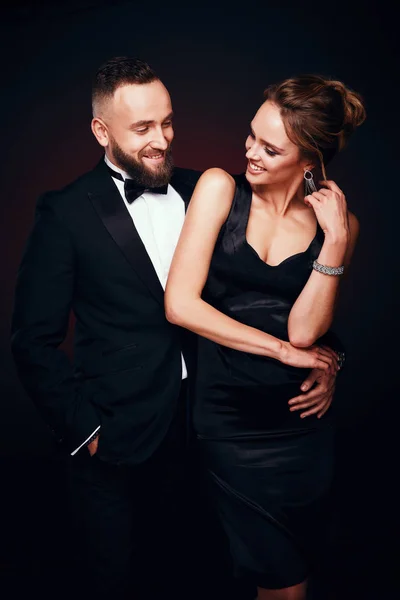 Chic, casal elegante: homem bonito com barba em smoking com mulher incrível com cabelo loiro updo, vestindo vestido preto sedoso e jóias de luxo, posando em estúdio escuro — Fotografia de Stock