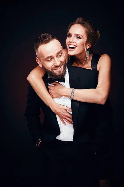 Casal luxuoso e elegante: homem barbudo bonito em smoking com mulher sorridente incrível com cabelo loiro updo, vestindo vestido preto sedoso e jóias chiques, posando em estúdio escuro — Fotografia de Stock