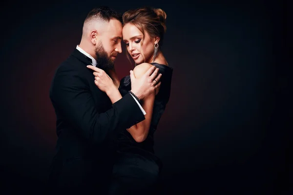 Sexy, luxury couple: handsome bearded man in tuxedo with pretty woman with blonde updo hair, wearing silky black dress and chic jewelry, posing in dark studio