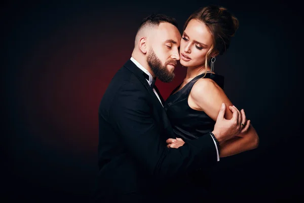 Pareja atractiva y elegante: hombre barbudo guapo en esmoquin con mujer increíble con cabello rubio, vestido negro sedoso y joyas elegantes, posando en un estudio oscuro — Foto de Stock