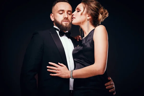 Casal elegante: homem barbudo bonito vestindo smoking com mulher incrível com cabelo loiro updo, vestindo vestido preto sedoso e jóias chiques, posando em estúdio escuro — Fotografia de Stock