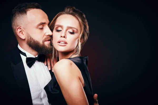 Sexy and luxury couple: handsome bearded man in tuxedo with amazing woman with blonde updo hair, wearing silky black dress and chic jewelry, posing in dark studio