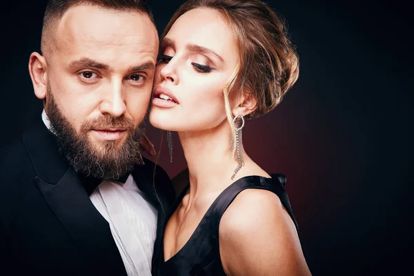 Casal luxuoso e elegante: homem barbudo bonito em smoking com mulher incrível com cabelo loiro updo, vestindo vestido preto sedoso e jóias chiques, posando em estúdio escuro — Fotografia de Stock