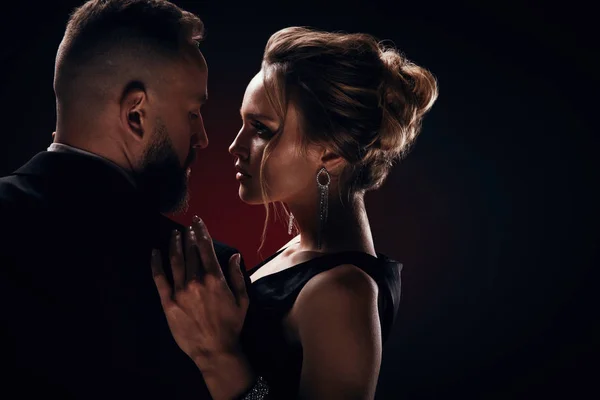 Passionné de luxe et bel homme barbu en smoking avec une femme étonnante avec des cheveux blonds updo, portant une robe noire soyeuse et des bijoux chics, posant dans un studio sombre — Photo