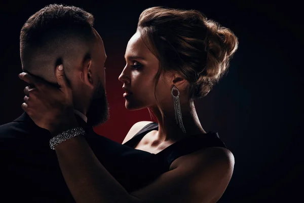 Tutkulu lüks lovers: yakışıklı adam smokin ile ipeksi siyah elbise ve şık takı, karanlık Studio'da poz giyen inanılmaz kadın sarışın kabarık saçlı sakallı