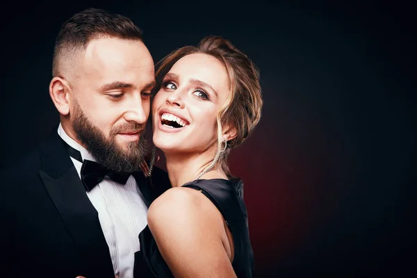 Casal elegante chique: homem barbudo bonito em smoking com mulher incrível com cabelo loiro updo, vestindo vestido preto sedoso e jóias chiques, posando em estúdio escuro — Fotografia de Stock