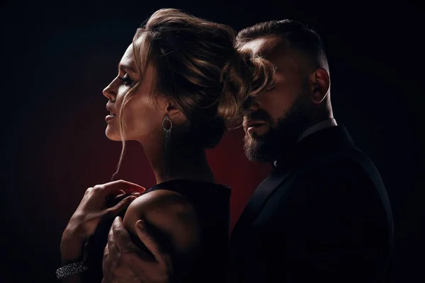 İpeksi siyah elbise ve şık takı ve yakışıklı sakallı adam ile harika bir smokin giyiyor lüks kadın sarışın kabarık saçlı koyu Studio'da poz