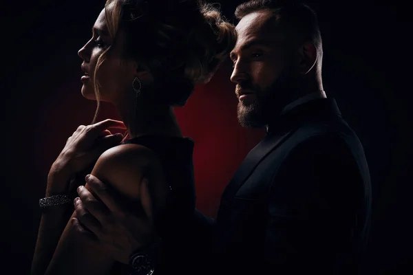 İpeksi siyah elbise ve şık takı ve yakışıklı sakallı adam ile harika bir smokin giyiyor tutkulu kadın sarışın kabarık saçlı koyu Studio'da poz