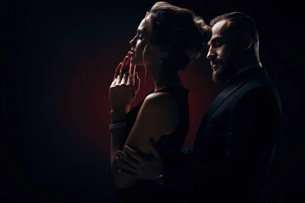 Şık çift: sarışın kabarık saçlı kadın ipeksi siyah elbise ve lüks mücevher ve yakışıklı sakallı adam ile harika bir smokin karanlık Studio'da poz giyiyor