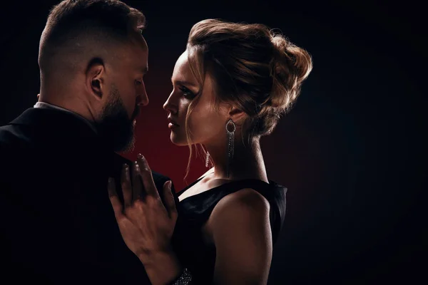 Passionate luxurious and handsome bearded man in tuxedo with amazing woman with blonde updo hair, wearing silky black dress and chic jewelry, posing in dark studio Stock Image