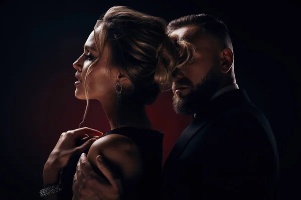Luxurious woman with blonde updo hair, wearing silky black dress and chic jewelry and handsome bearded man in tuxedo with amazing, posing in dark studio Royalty Free Stock Photos