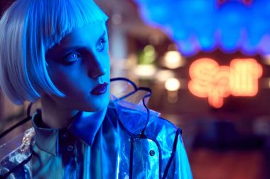 Fütüristik neon ışıklı ipeksi bluz ve şeffaf yağmurluk giymiş beyaz bob saç modeli ile seksi bir kadın portresi kapatın
