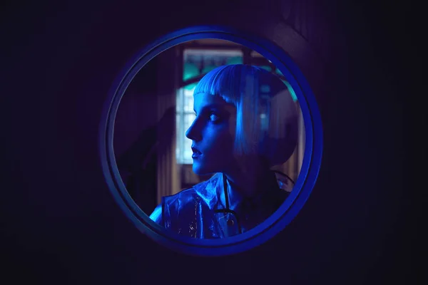 Futuristic neon lighted portrait of amazing woman with white bob hairstyle wearing silky blouse and transparent raincoat at the round window — Stock Photo, Image