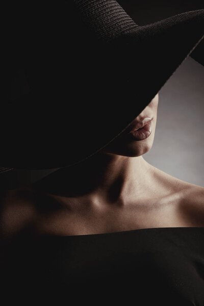 Dramatic studio portrait of elegant woman in black wide hat and black dress. Hidden half face.