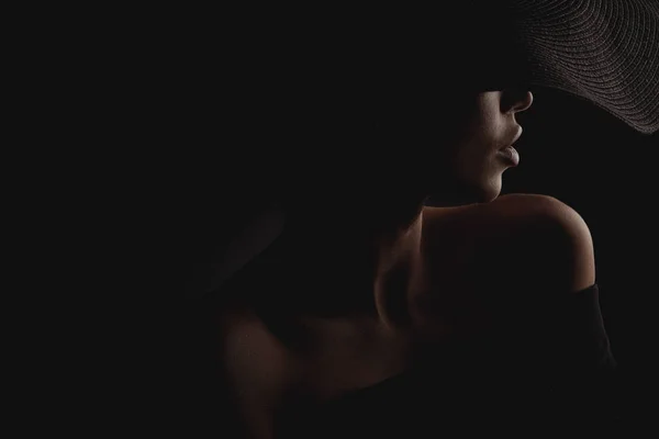 Dramatic dark studio portrait of elegant and sexy woman in black wide hat and black dress. Hidden half face.
