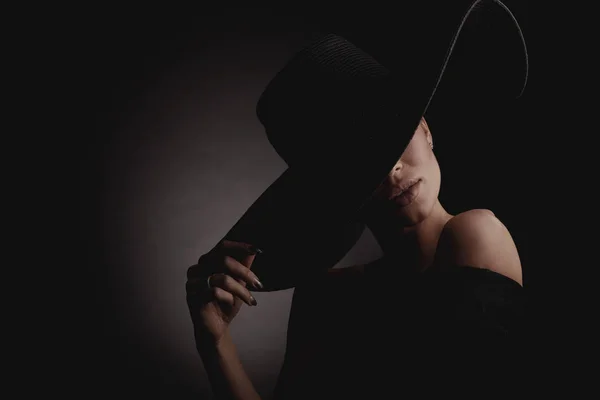 黒の広い帽子と黒のドレスでエレガントな女性の劇的な暗いスタジオ肖像画 隠された目 — ストック写真