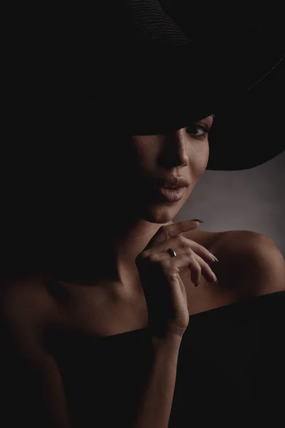 Dark studio portrait of elegant and sexy young woman in black wide hat and black dress.