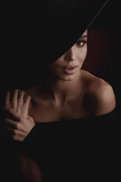 黒の広い帽子と黒のドレスでエレガントでセクシーな若い女性の暗いスタジオ ポートレート — ストック写真