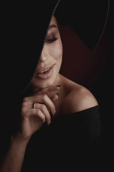 黒の広い帽子と黒のドレスでエレガントでセクシーな笑顔の女性の劇的な暗いスタジオ ポートレート — ストック写真