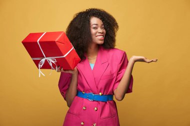 Afro saç modeli ile çekici genç Afrikalı kız Güzellik Portresi. Pembe ceket giyen kız gülümseyerek, mavi tozluk ve kemer tutan elini ve Sarı zemin üzerine poz bir hediye. Stüdyo vurdu