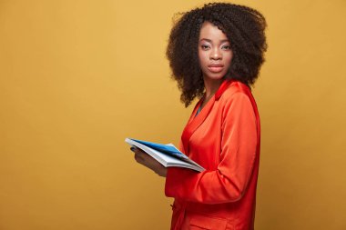 Renkli afro saç modeli ile genç Afrikalı kız portresi. Turuncu ceket giyen ciddi kız onun elinde boşaltmak ve poz sarı arka plan üzerinde tutar. Stüdyo vurdu.