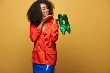Parlak ve renkli afro saçlı genç ve güzel Afrikalı kız portresi. Turuncu ceket giyen gülümseyen kız yeşil ayakkabı parmaklarını üzerinde tutar. Stüdyo vurdu