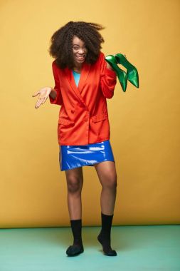 Genç ve güzel Afrikalı kız afro saçlı renkli portresi. İpeksi turuncu ceket, Menekşe Lateks etek ve siyah çorap giyen gülümseyen kız yeşil ayakkabı onun elinde tutar. Stüdyo vurdu