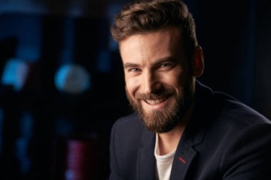 Koyu saç, sakal ve bıyık koyu ceket giyen genç gülümseyen yakışıklı portresi. Stüdyo ile Mavi ışıklı tuğla arka plan bulanık saat ve kırmızı varil ile vurdu