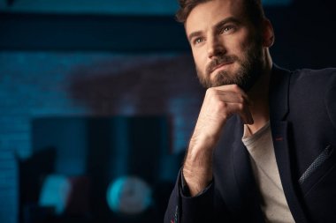 Koyu saç, sakal ve bıyık koyu ceket dalgın yakışıklı adam portresi. El yüz yakın. Stüdyo ile Mavi ışıklı tuğla arka plan bulanık saat ve kırmızı varil ile vurdu
