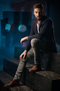 Stüdyo portresi ciddi yakışıklı adam koyu saç, sakal ve bıyık koyu mavi ceket, gri pantolon ve ahşap yüzey üzerinde oturan kahverengi ayakkabı giyiyor. Saat ve varil ile Mavi ışıklı tuğla arka plan