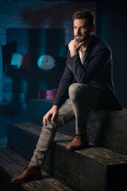 Stüdyo portre çekici yakışıklı adam koyu saç, sakal ve bıyık koyu mavi ceket, gri pantolon ve ahşap merdivenlerde oturan kahverengi ayakkabı giyiyor. Saat ve varil ile Mavi ışıklı tuğla arka plan