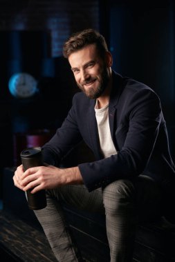 Stüdyo portre çekici gülümseyen adam koyu saç, sakal ve bıyık koyu mavi ceket, gri pantolon ve ellerde termo kupa ile ahşap yüzey üzerinde oturan kahverengi ayakkabı giyiyor. Saat ve varil ile Mavi ışıklı tuğla arka plan