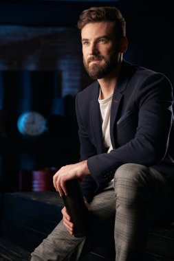 Stüdyo portresi ciddi adam koyu saç, sakal ve bıyık koyu mavi ceket, gri pantolon ve ellerde termo kupa ile ahşap yüzey üzerinde oturan kahverengi ayakkabı giyiyor. Saat ve varil ile Mavi ışıklı tuğla arka plan