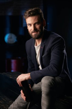 Stüdyo portre çekici adamla koyu saç, sakal ve bıyık koyu mavi ceket, gri pantolon ve ellerde termo kupa ile ahşap yüzey üzerinde oturan kahverengi ayakkabı giyiyor. Saat ve varil ile Mavi ışıklı tuğla arka plan