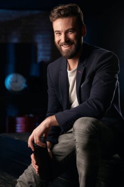 Koyu saç, sakal ve bıyık koyu mavi ceket, gri pantolon ve ellerde termo kupa ile ahşap yüzey üzerinde oturan kahverengi ayakkabı giyen adamla gülümseyen studio portresi. Saat ve varil ile Mavi ışıklı tuğla arka plan