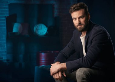Stüdyo portresi ciddi yakışıklı adam koyu saç, sakal ve bıyık koyu mavi ceket, gri pantolon giyiyor. Saat ve varil ile Mavi ışıklı tuğla arka plan