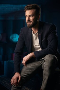 Stüdyo portresi ciddi yakışıklı adam koyu saç, sakal ve bıyık koyu mavi ceket, gri pantolon ahşap yüzey üzerinde oturan giyiyor. Saat ve varil ile Mavi ışıklı tuğla arka plan