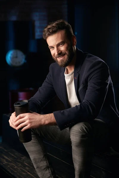 Studio portrait of attractive smiling man with dark hair, beard and mustache wearing dark blue jacket, gray pants and brown shoes, sitting on wooden surface with termo mug in hands. Blue lighted brick background with clock and barrel