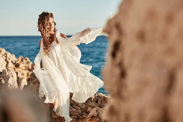 美丽性感的年轻女性模型与一头乌黑的长发 戴着长长的白色礼服在夏天在暗蓝色海附近的岩石上构成的肖像 — 图库照片