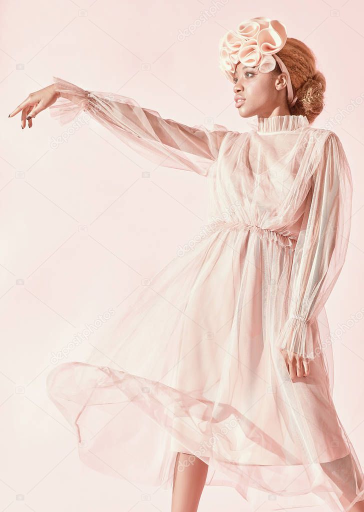 Studio fashion portrait of beautiful black woman with light hair and green eyes wearing powder pink tulle dress and big frill hairband and posing. Pink background