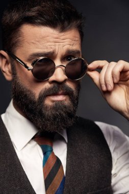Koyu saç, bıyık ve sakal beyaz gömlek, kahverengi yelek ve renkli kravat alır yuvarlak güneş gözlüğü kapalı ciddi yakışıklı adamla düşük anahtar portresi