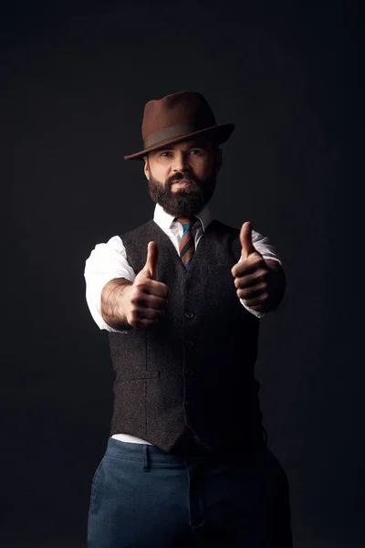 Studio portrait of handsome man with dark hair and mustache in white shirt, brown vest, colorful tie, brown hat with thumbs up. Old fashioned style, gentleman.