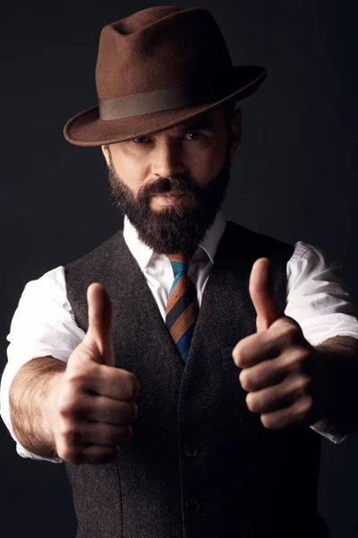 Studio portrait of serious handsome man with dark hair and mustache in white shirt, brown vest, colorful tie and brown hat with thumbs up. Old fashioned style, gentleman.
