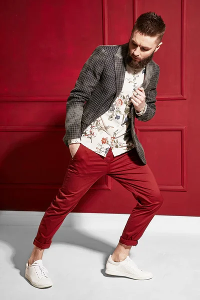 黒い髪と赤いズボンを身に着けているひげでハンサムな男のファッションの肖像画 花柄のシャツ 格子縞のジャケット 赤の背景 — ストック写真