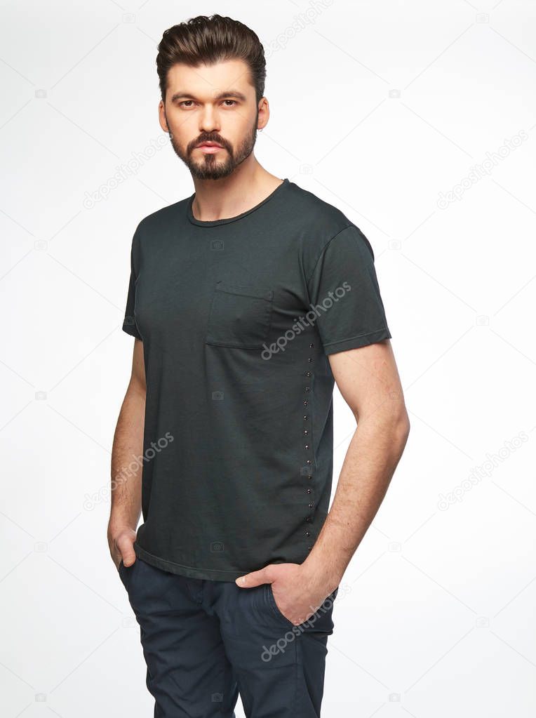 Fashion portrait of handsome male model with dark hair, beard and eyes, wearing grey t-shirt and pants and posing on white background