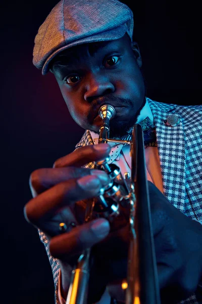 Neon studio portrait of handsome black man in plaid jacket and vintage cap playing trumpet. Orange and blue light