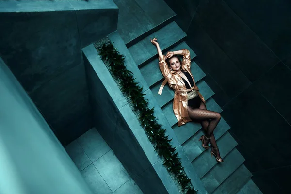 Amazing young woman in gold trench coat, black bodysuit with gold belt and in black polka dot tights is lying on the turquoise stairs